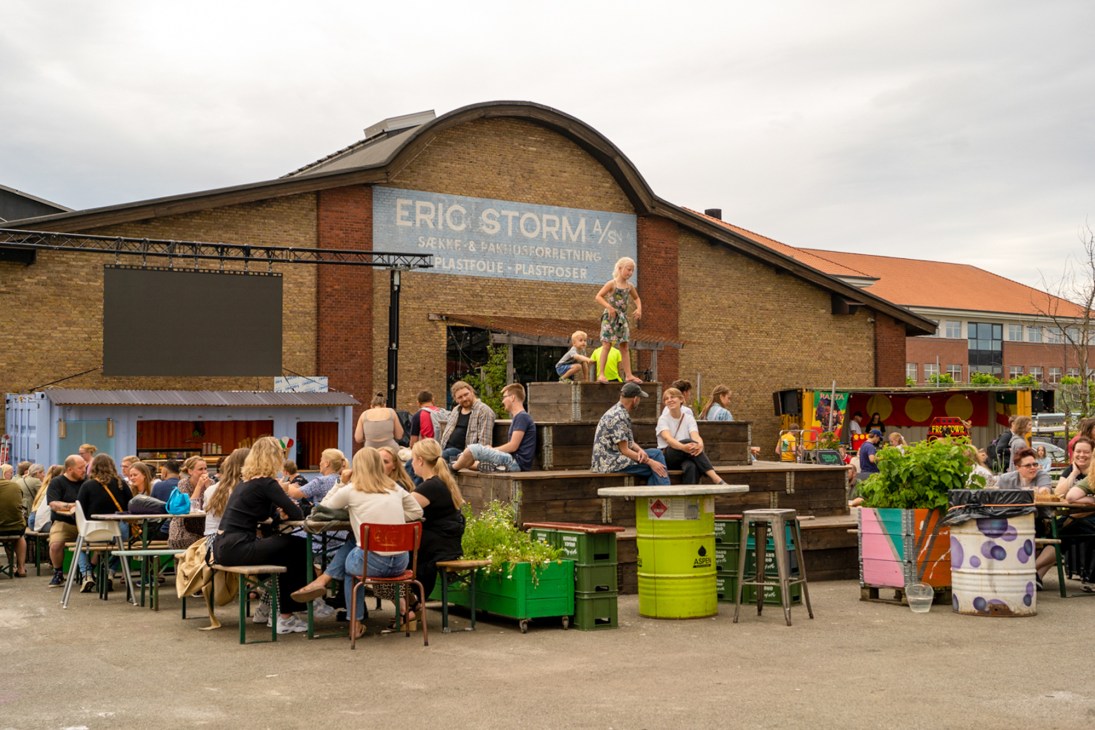 People dining at outdoor tables in front of brick warehouse building with Eric Storm sign in Denmark