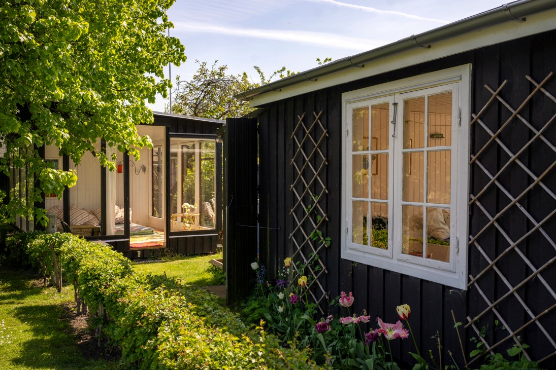 Black wooden house with white-framed windows and lattice trellis, surrounded by garden beds with colorful flowers