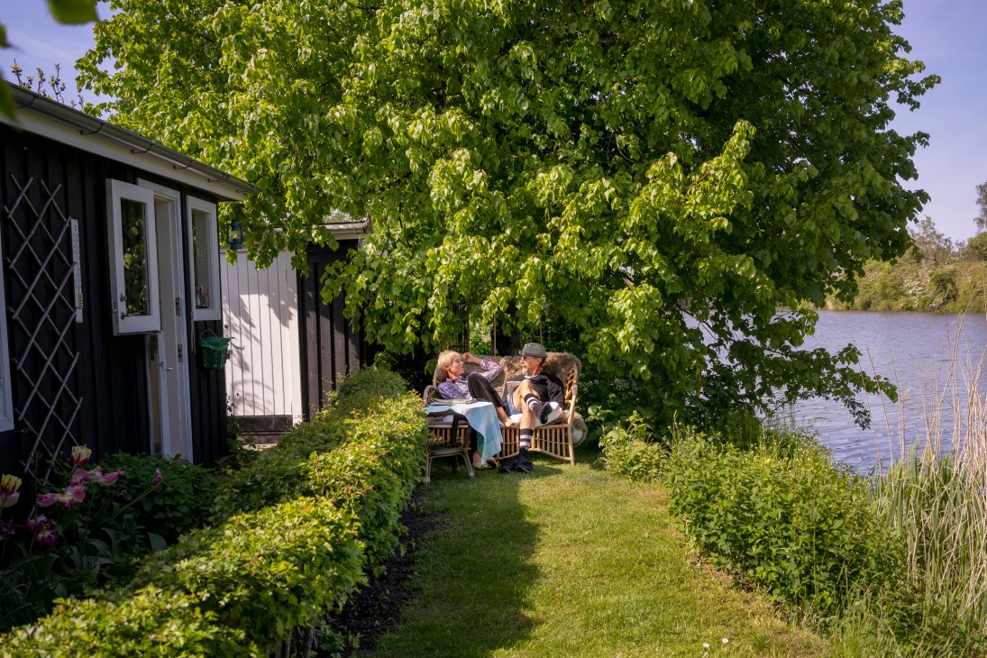 Signe Sejlund and Hans Bullitt Fogh relaxing in wooden chairs under a large tree by water at their kolonihave