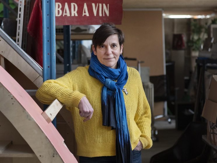 Woman in yellow sweater and blue scarf standing by stairs in warehouse with "BAR A VIN" sign overhead