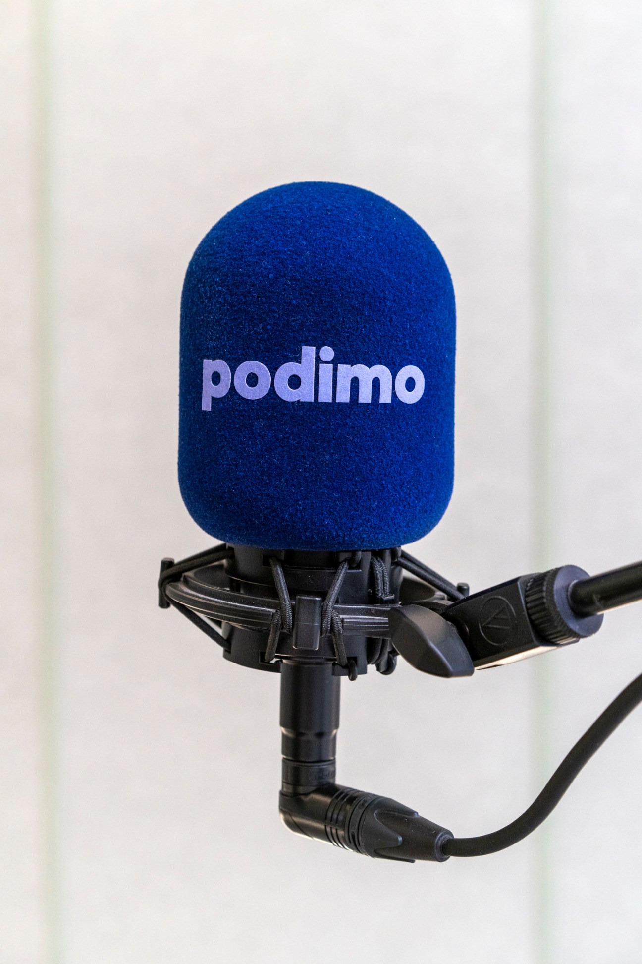Podimo-branded microphone with blue foam windscreen mounted on black shock mount.
