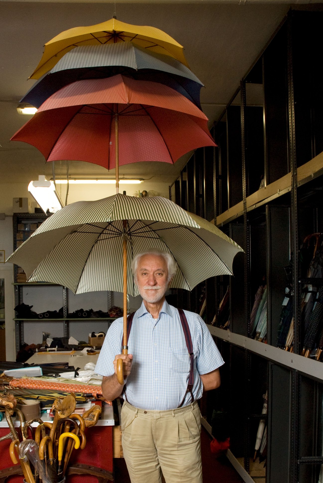 Elderly man holding striped umbrella in workshop with colorful umbrellas hanging from ceiling at Ombrelli Maglia