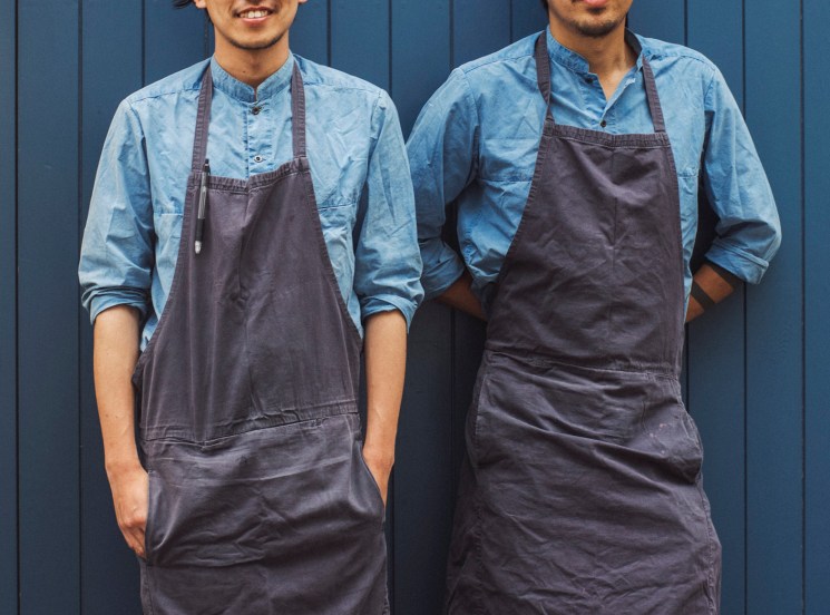 Two people in matching blue shirts and dark aprons stand side by side against a navy wall.