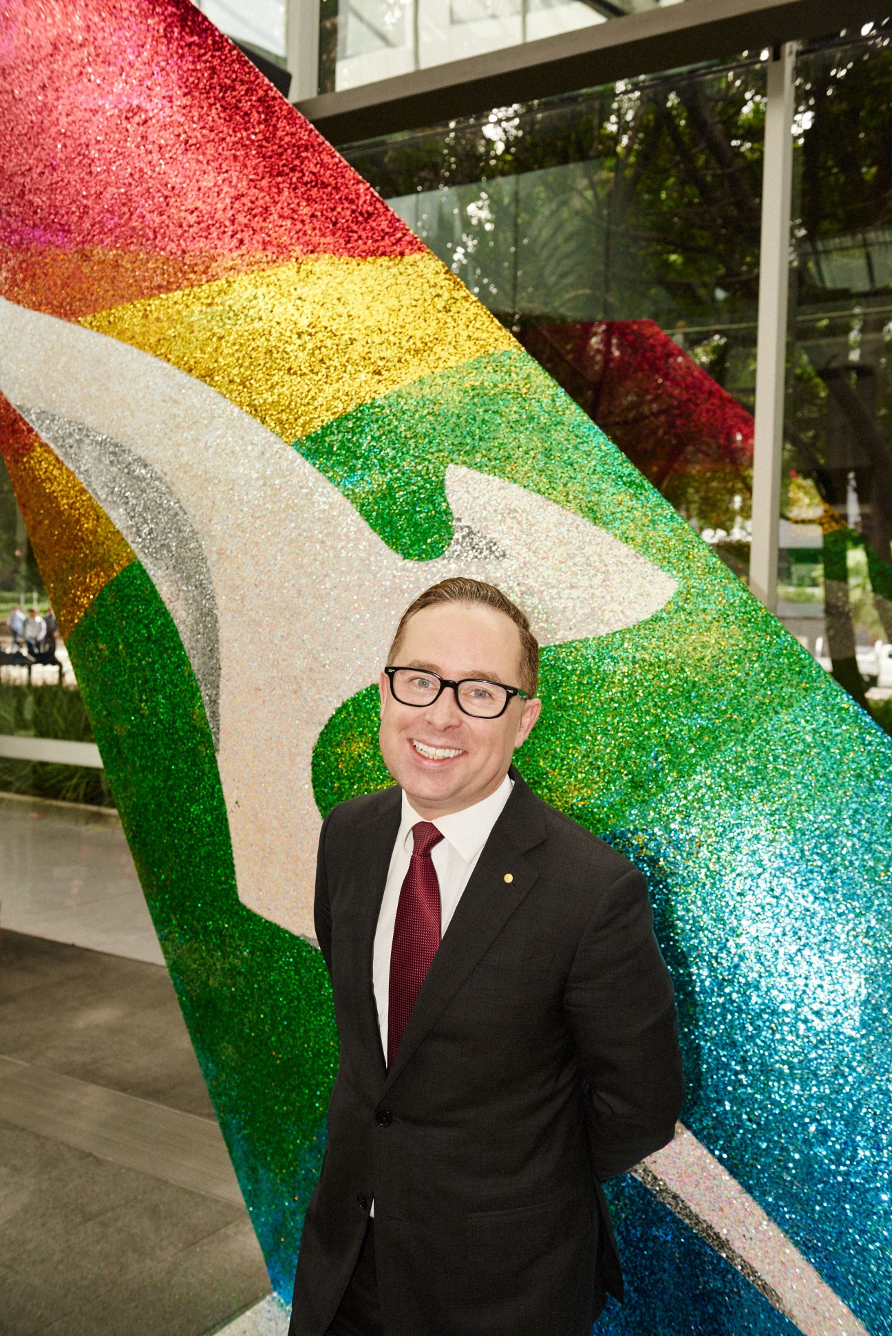 Alan Joyce, Qantas CEO, stands in front of a colorful geometric art installation with rainbow-colored glittering panels.