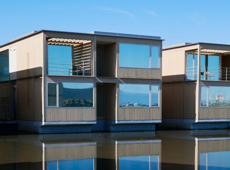 Modern two-storey floating homes with large glass windows and roof terraces reflected on calm water.