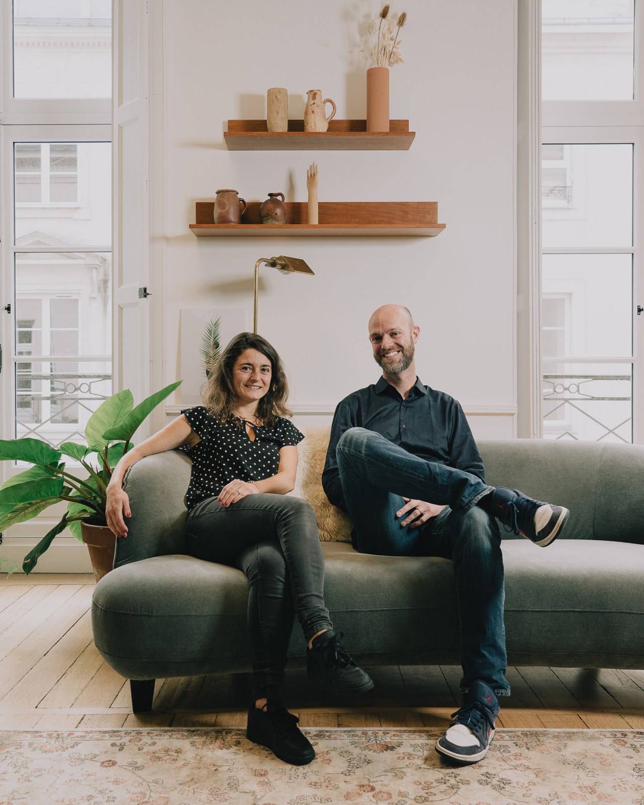 Nolwenn Belléguic and Matthieu Guesné, co-founders of Lhyfe, sit on a gray sofa in a bright living room with shelved ceramics and dried flowers behind them.