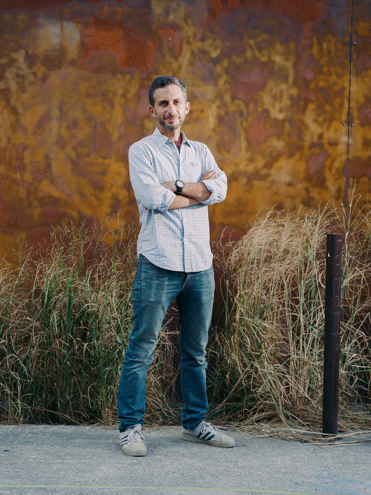 Rob Spiro, founder of Imagination Machine, standing with arms crossed in front of a weathered rust-coloured wall with dry grasses.