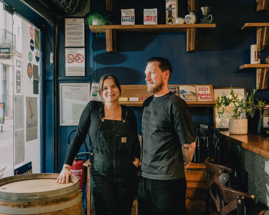 Sarah Mainguy and Damien Crémois stand in their restaurant Vacarme, with dark blue walls, wooden shelves, and bar setup behind them.