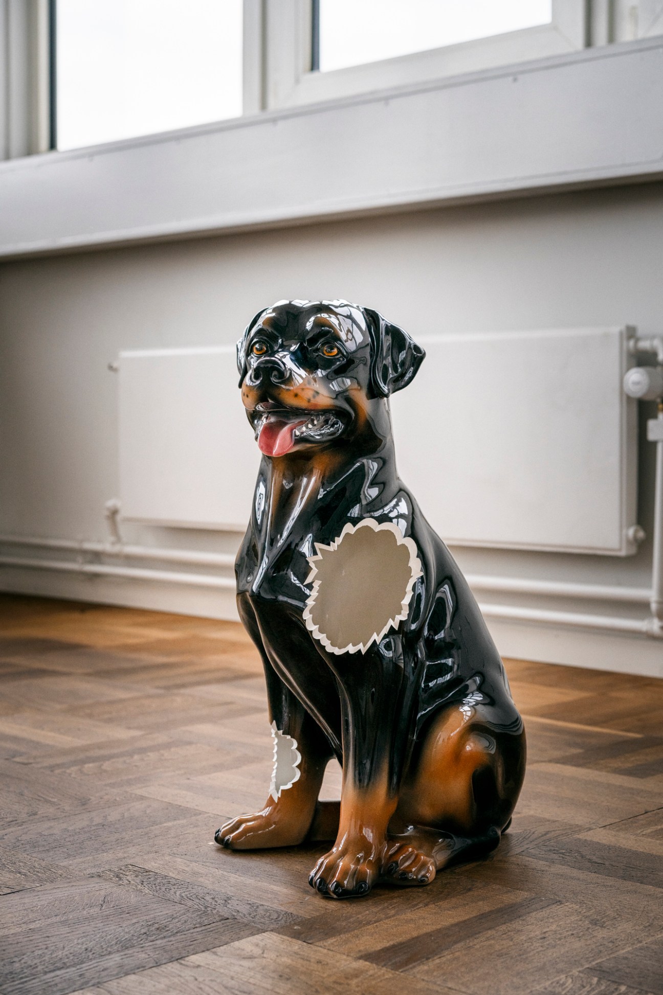 Black ceramic dog sculpture with tan markings sitting on wooden floor in gallery space