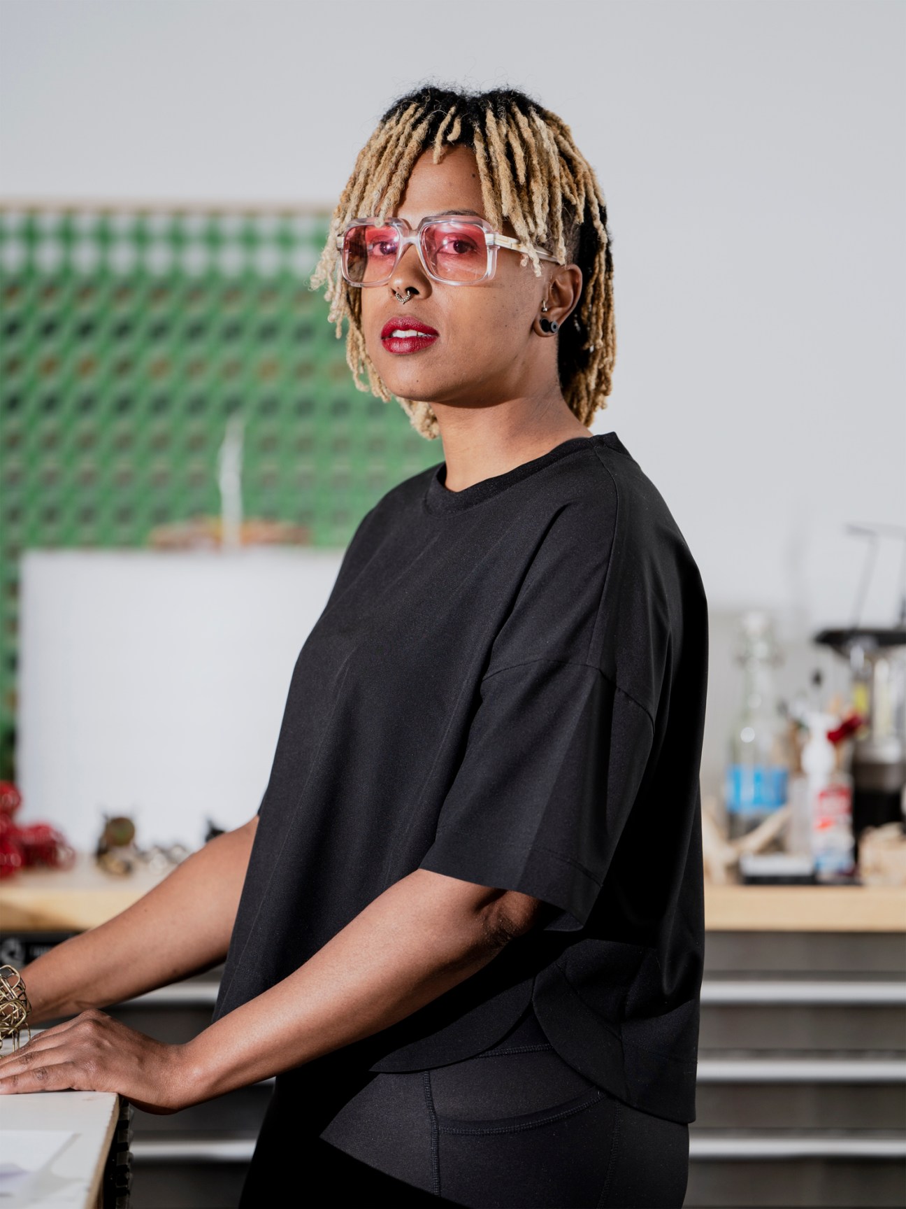 Person in black t-shirt and pink-tinted glasses stands in a kitchen or studio space.