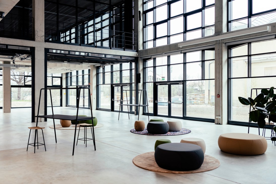 Modern office space with floor-to-ceiling windows, concrete pillars, and casual seating with round cushions and wooden tables.