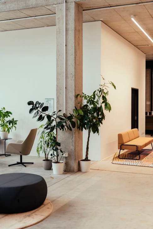 Modern office space with concrete pillars, potted plants, tan chair, and orange bench seating in natural light.