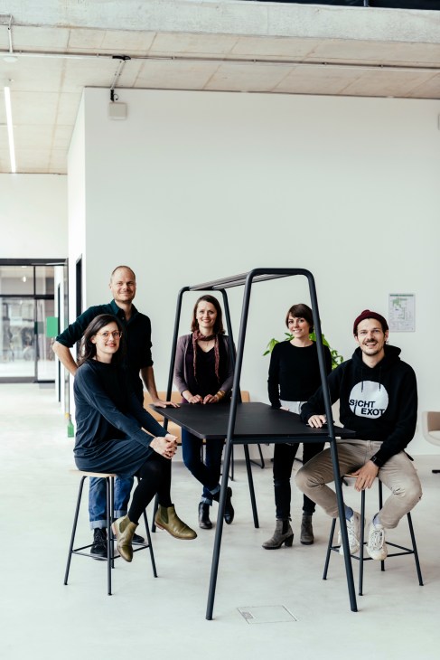 Five team members of Sichtbarmacher pose together on black chairs in a bright, minimalist office space with white walls and exposed ceiling.