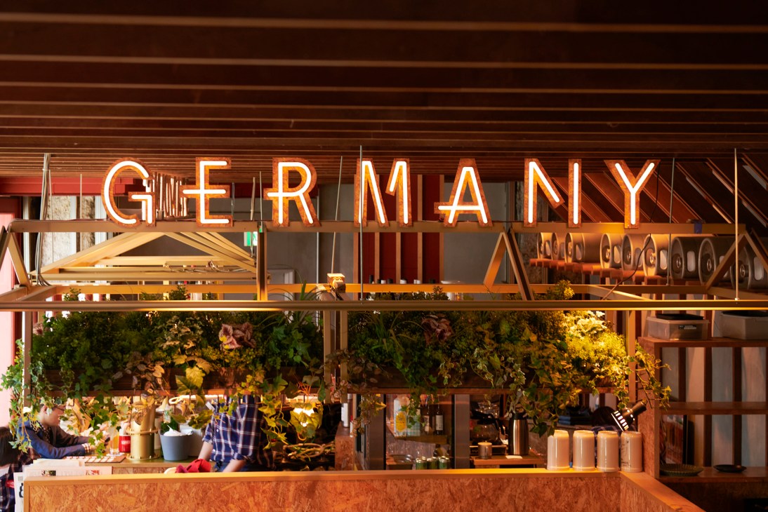 Modern restaurant interior with neon 'GERMANY' sign, open kitchen, greenery wall, and contemporary design