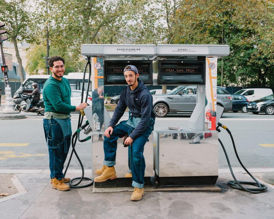 Two young men posing at a modern fuel pump station in Paris, one holding a fuel nozzle