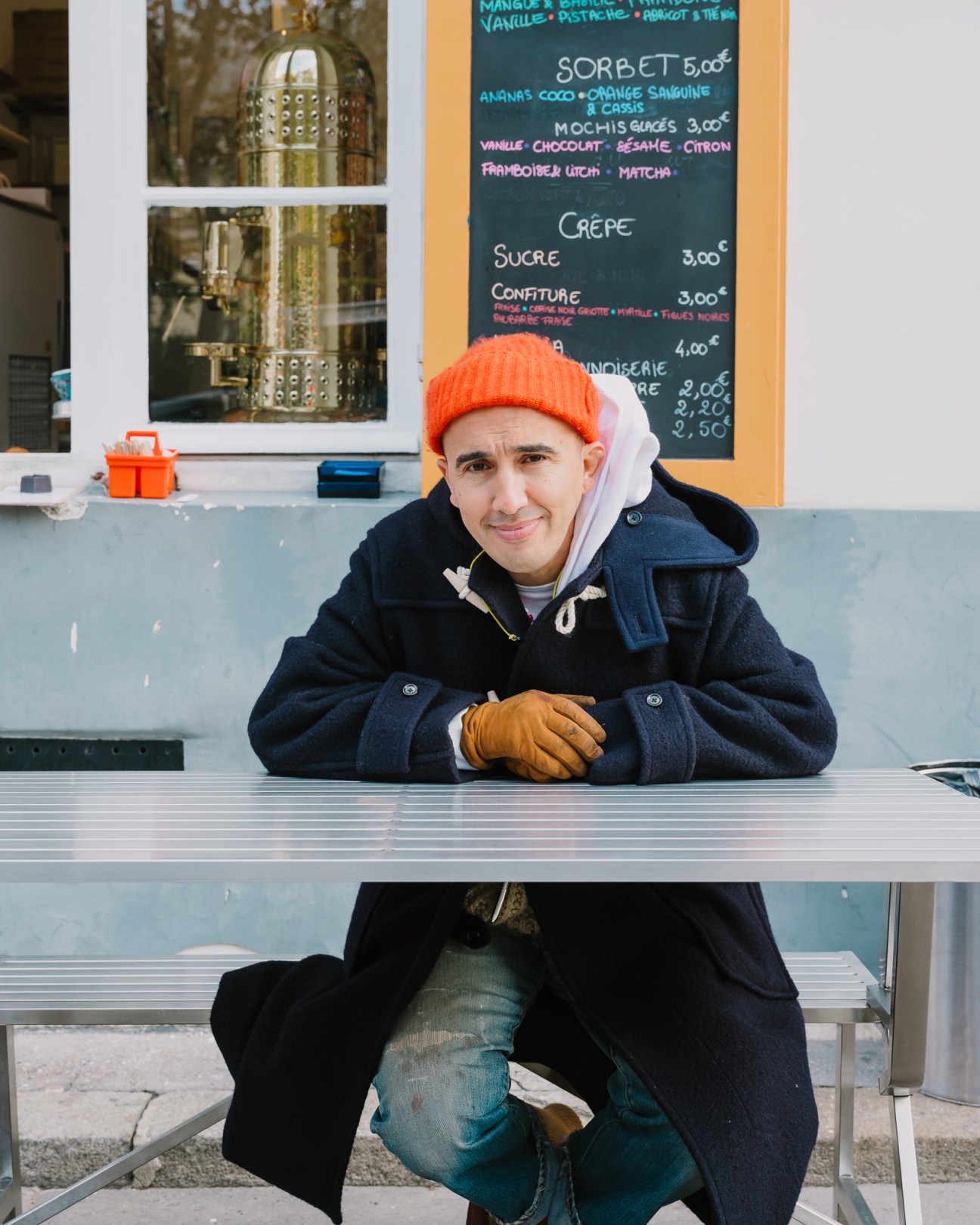 Ramdane Touhami in orange beanie and dark coat sitting at counter inside cafe with menu board visible