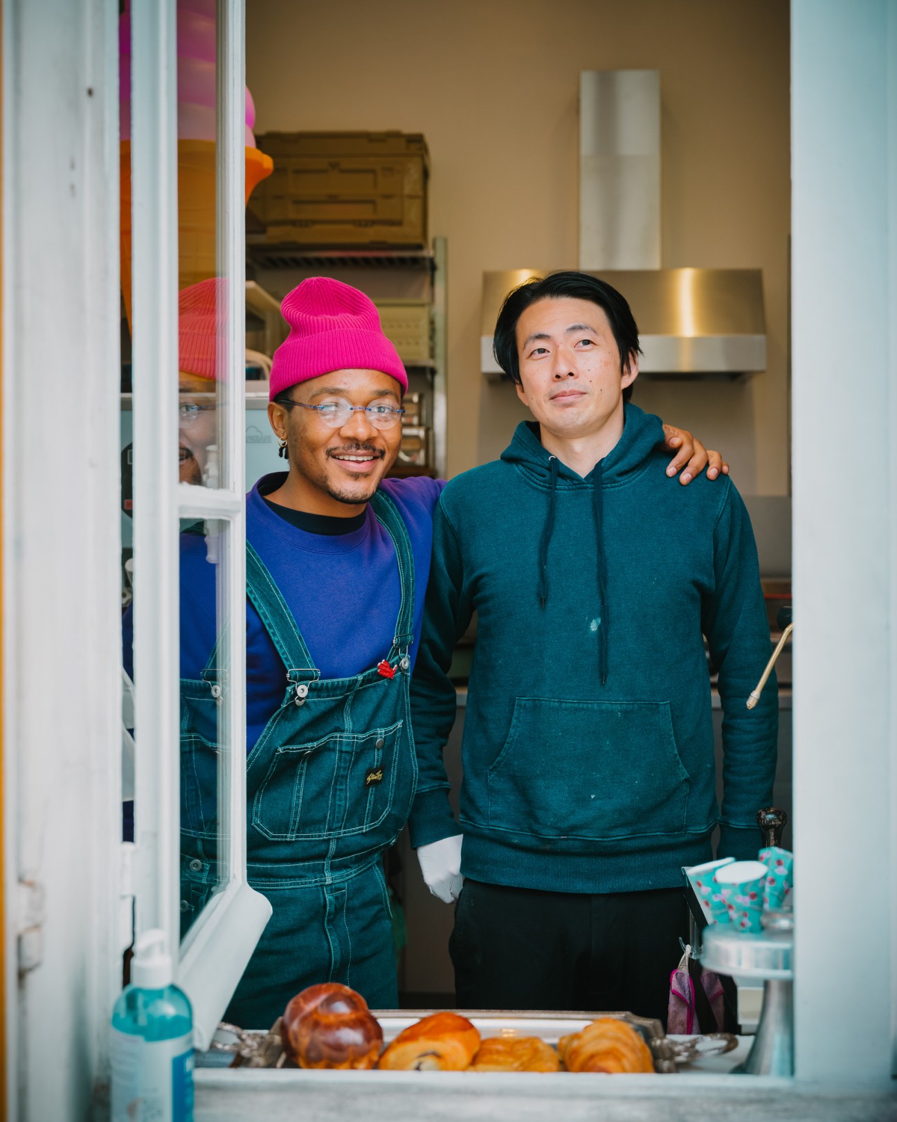 Two men standing together in a service station, one wearing a pink beanie and overalls, the other in a green hoodie