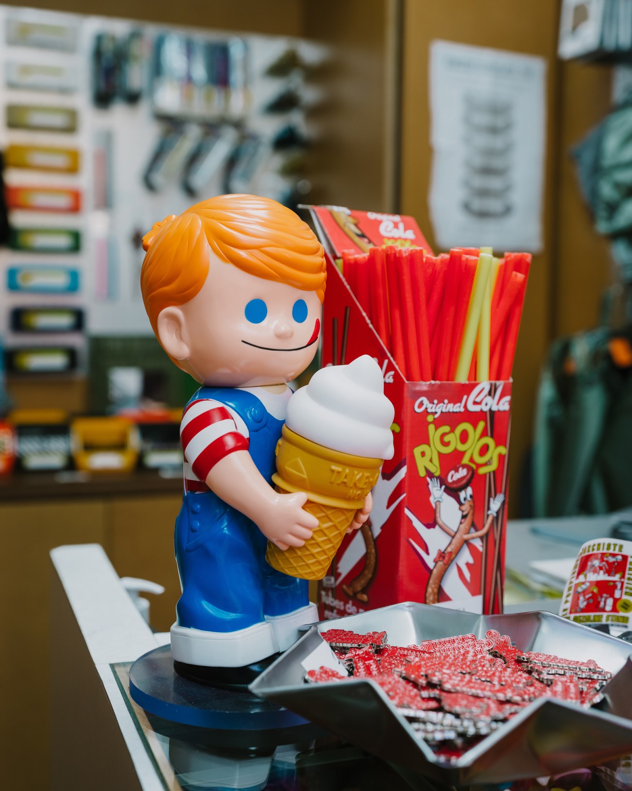Plastic figurine of blonde boy in blue overalls holding ice cream cone next to red straws and candy packages