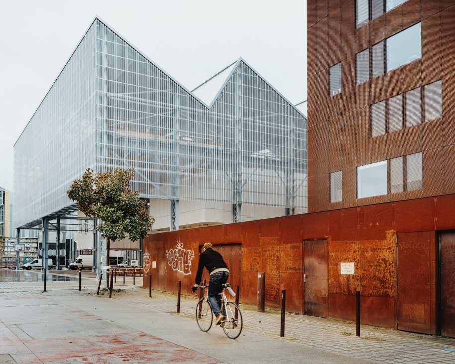Modern glass building with peaked roof next to brick structure, cyclist riding past on street