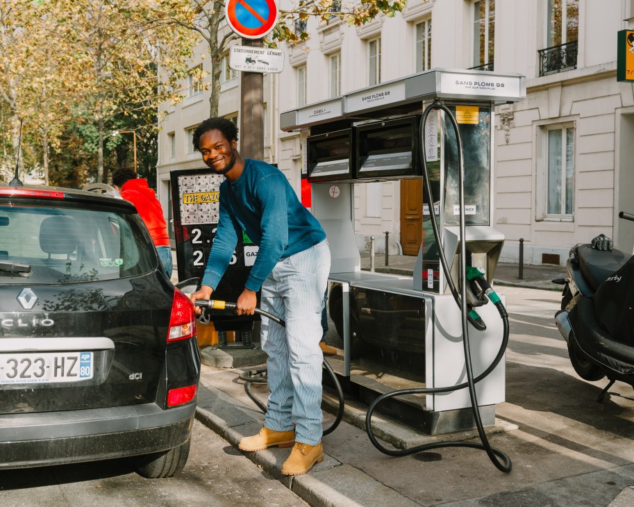 A smiling man in a blue sweater and gray pants fills up a black Renault Clio at a Parisian gas station