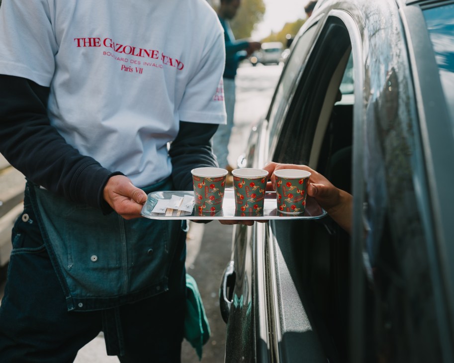 Person in white t-shirt holding metal tray with three patterned paper coffee cups next to car