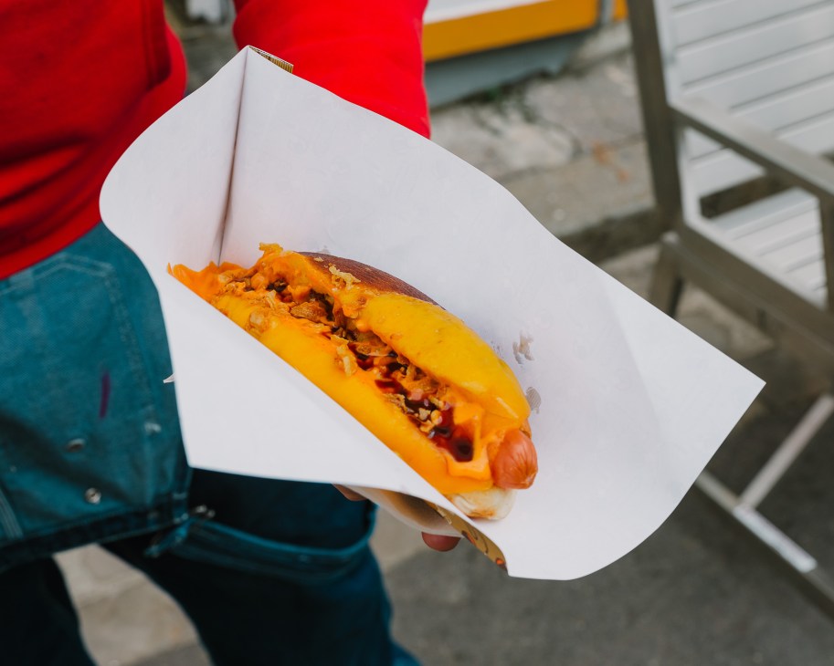 Person in red jacket and blue jeans holding a hotdog in white paper wrapper outdoors