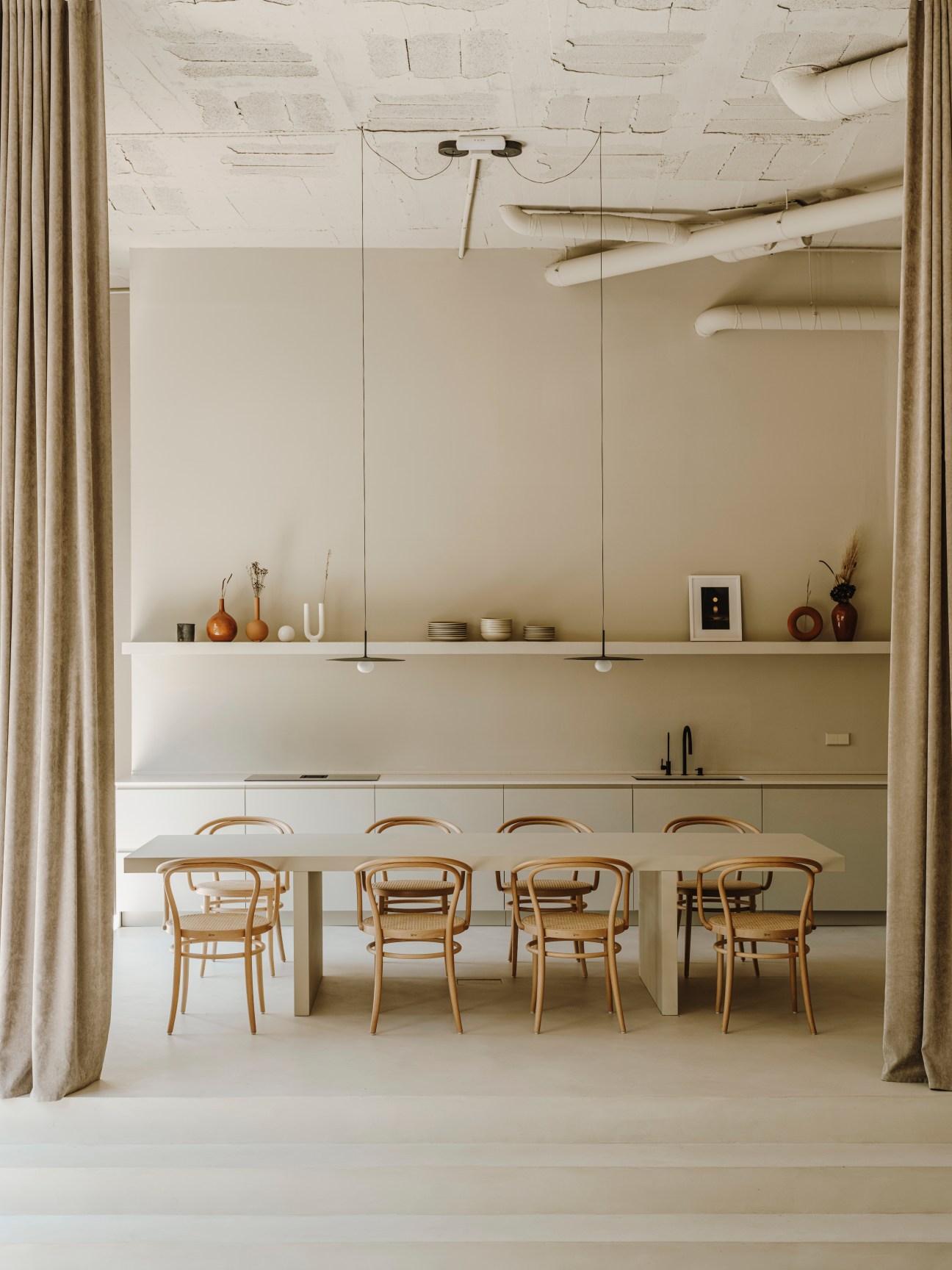 Minimalist dining room with a long light table and bentwood chairs, a floating shelf with decorative vases and objects above, and neutral tones throughout.