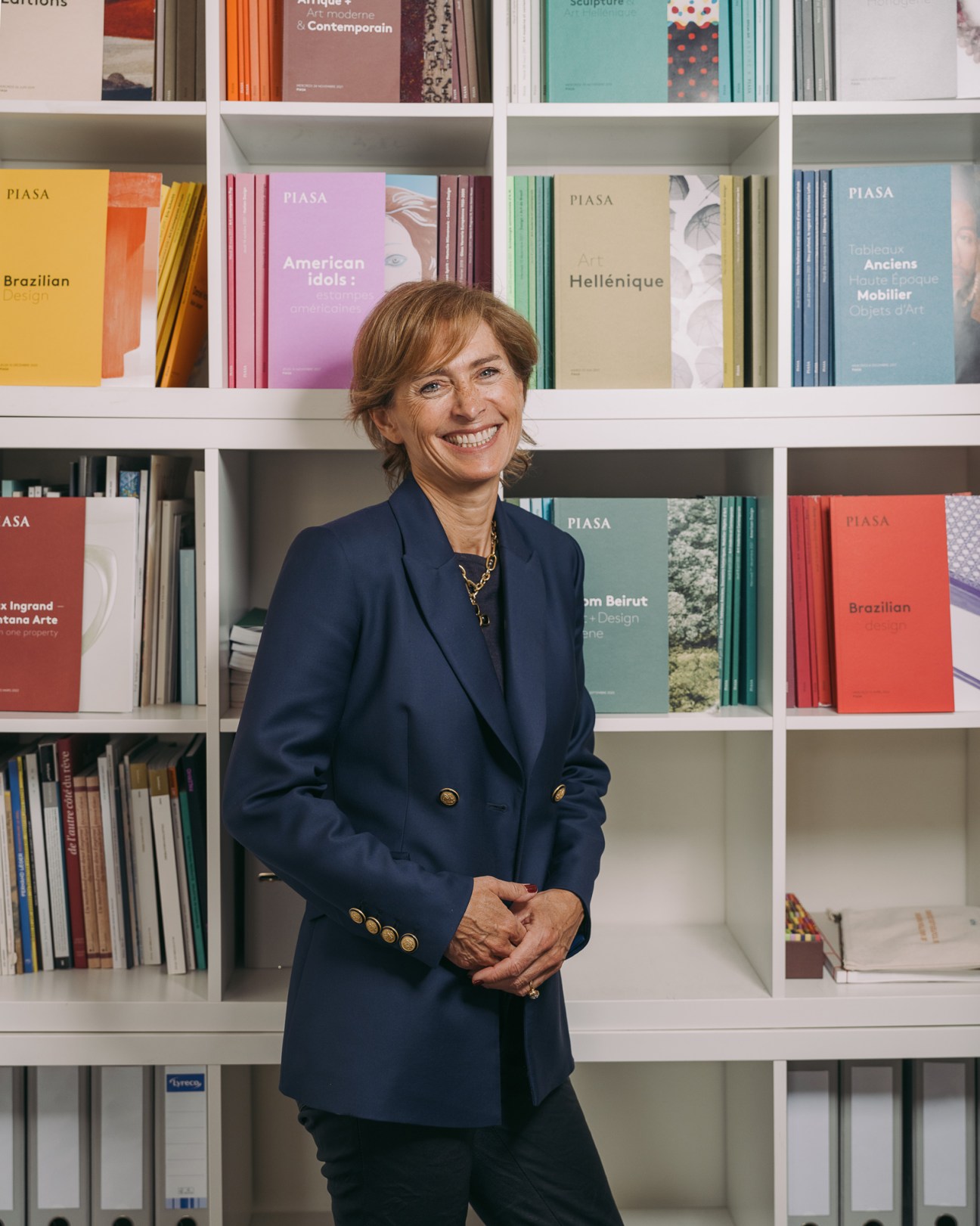 Marie Filippi, CEO of Piasa auction house, standing in front of colorful catalogues on shelves.