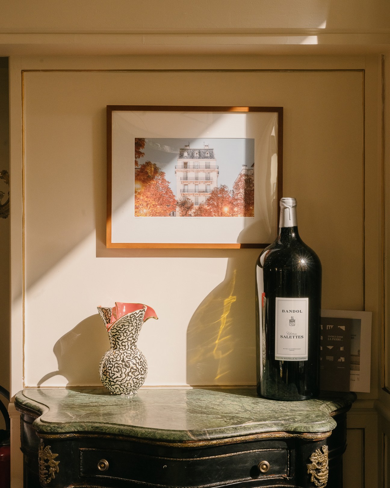 Styled interior vignette with framed Parisian building photograph, patterned ceramic vessel, and wine bottle on marble table.