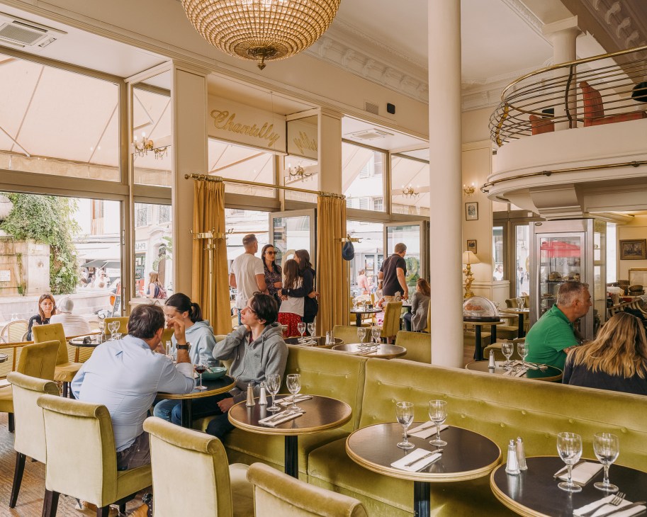 Elegant café interior with ochre velvet banquette seating, beige ceiling with decorative pendant light, and large windows overlooking a European street.