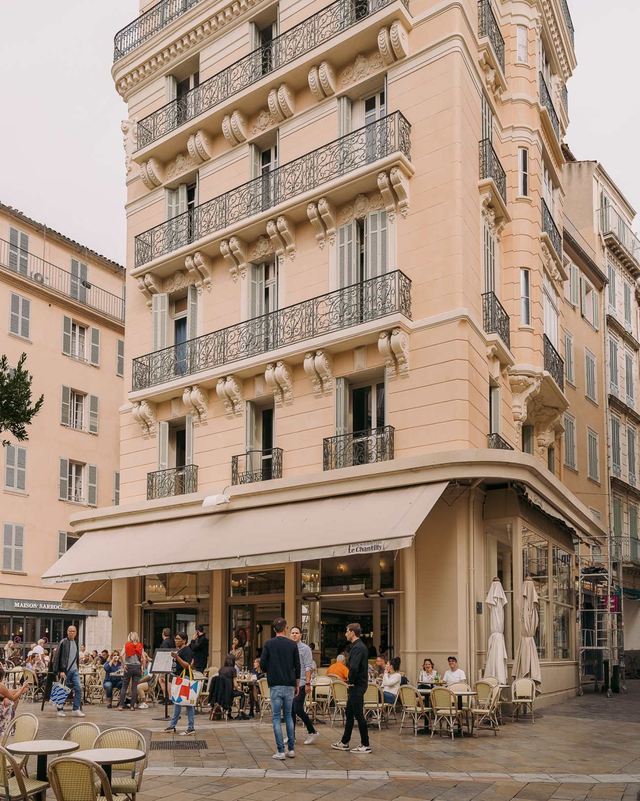 Peach-colored Haussmann-style building with ornate stonework and wrought-iron balconies overlooking a plaza with café seating and pedestrians.