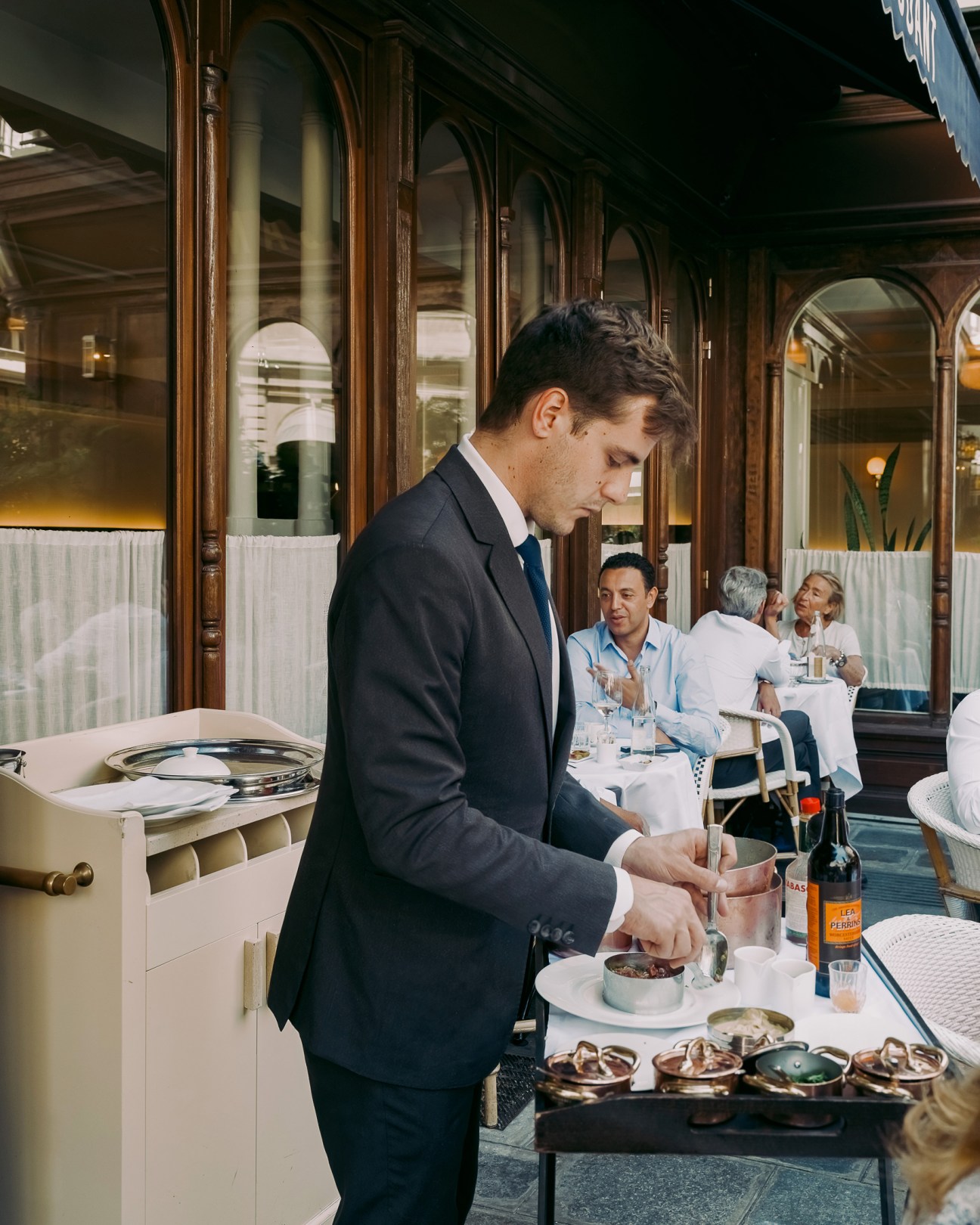A waiter in a dark suit serves food from a trolley in an elegant restaurant with ornate windows and diners seated nearby