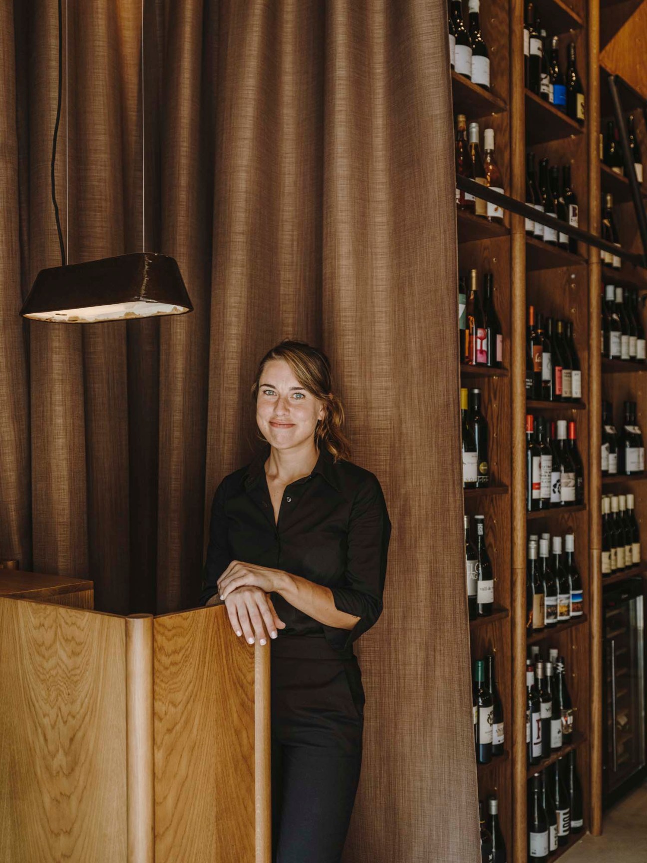 A woman in black uniform stands smiling beside wooden wine storage shelves in an upscale restaurant interior