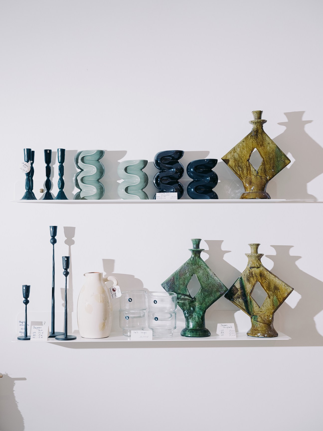 Ceramic vessels and candle holders displayed on white shelving, including wavy forms in blue and cream, dark sculptural pieces, and a gold diamond-shaped object.