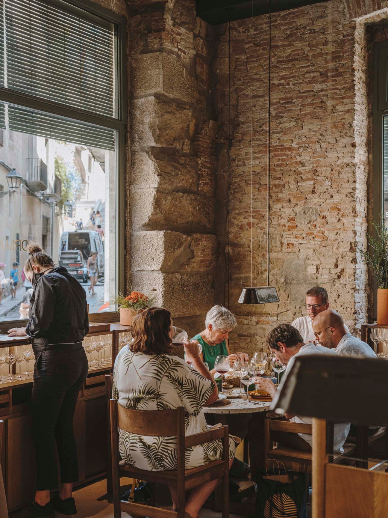 Diners enjoy wine and conversation at wooden tables in a rustic restaurant with exposed stone walls and large windows