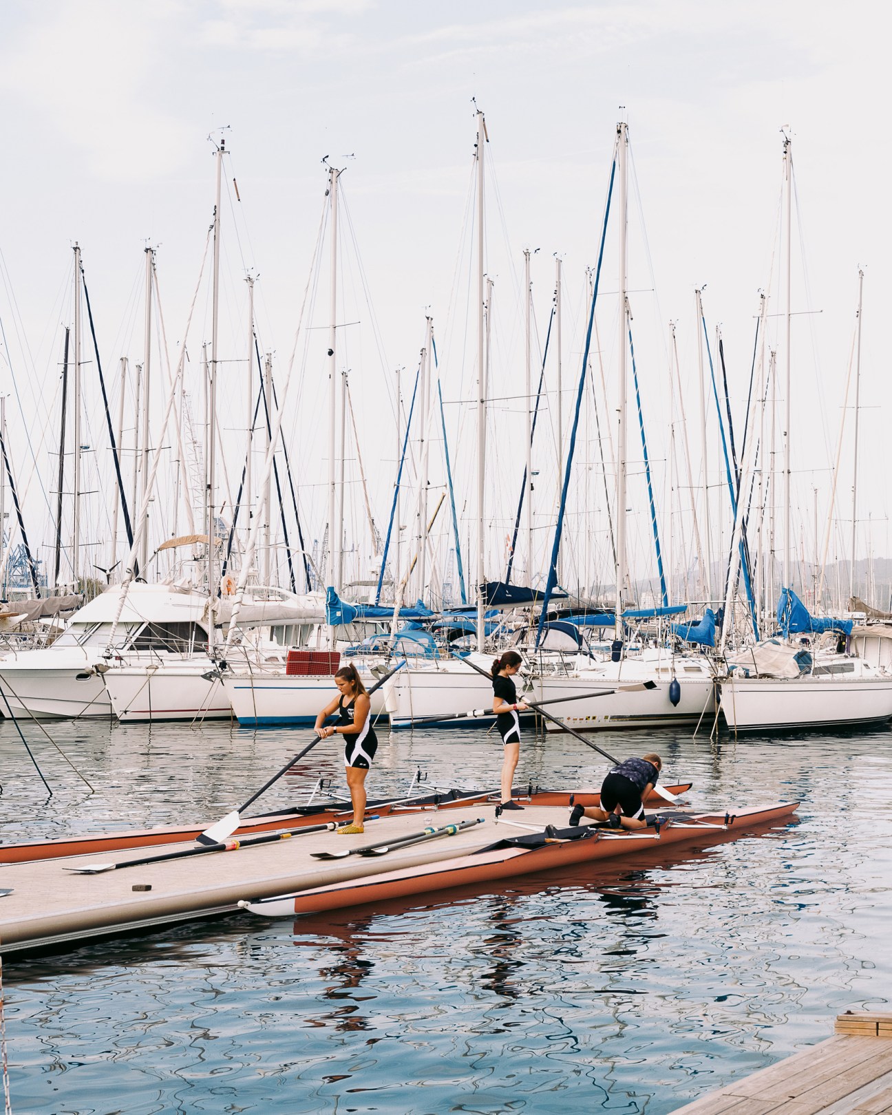 Three people prepare stand-up paddleboards in water with moored sailboats in background.
