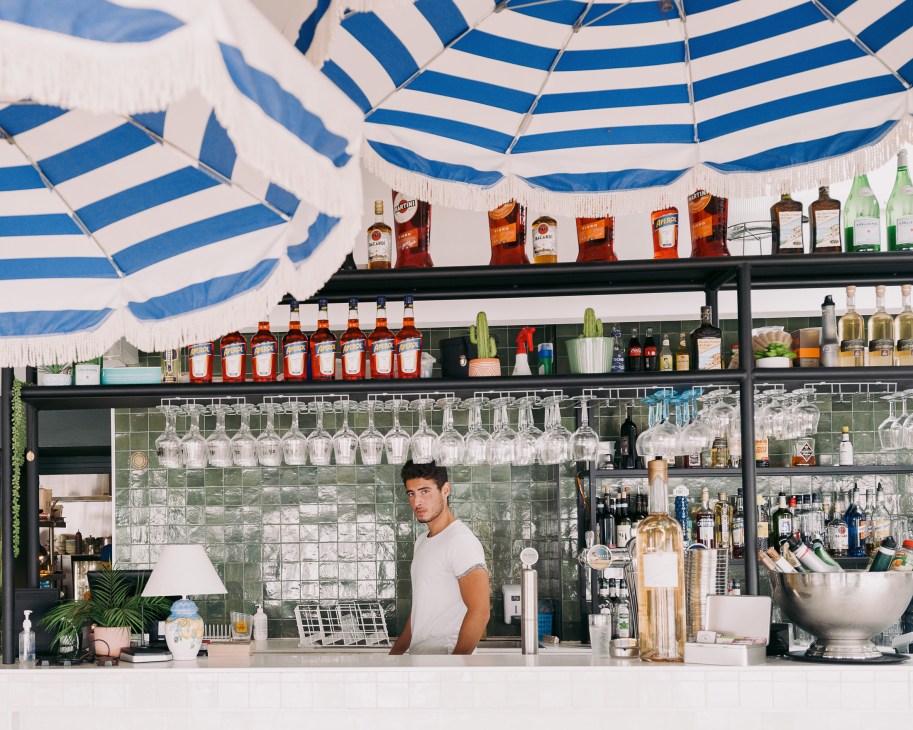 Bartender in white shirt standing behind bar with blue and white striped ceiling and shelves of spirits