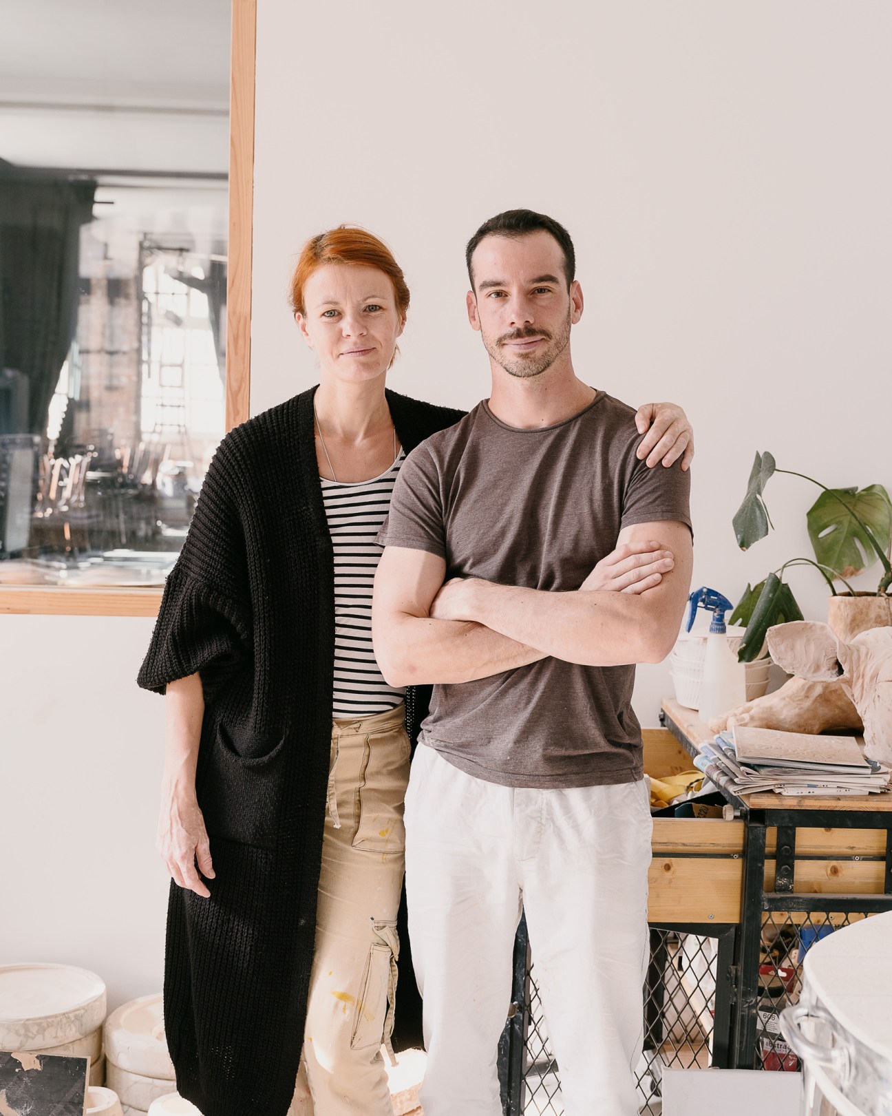 Two people stand together in a bright studio space with a potted plant and shelving visible behind them.