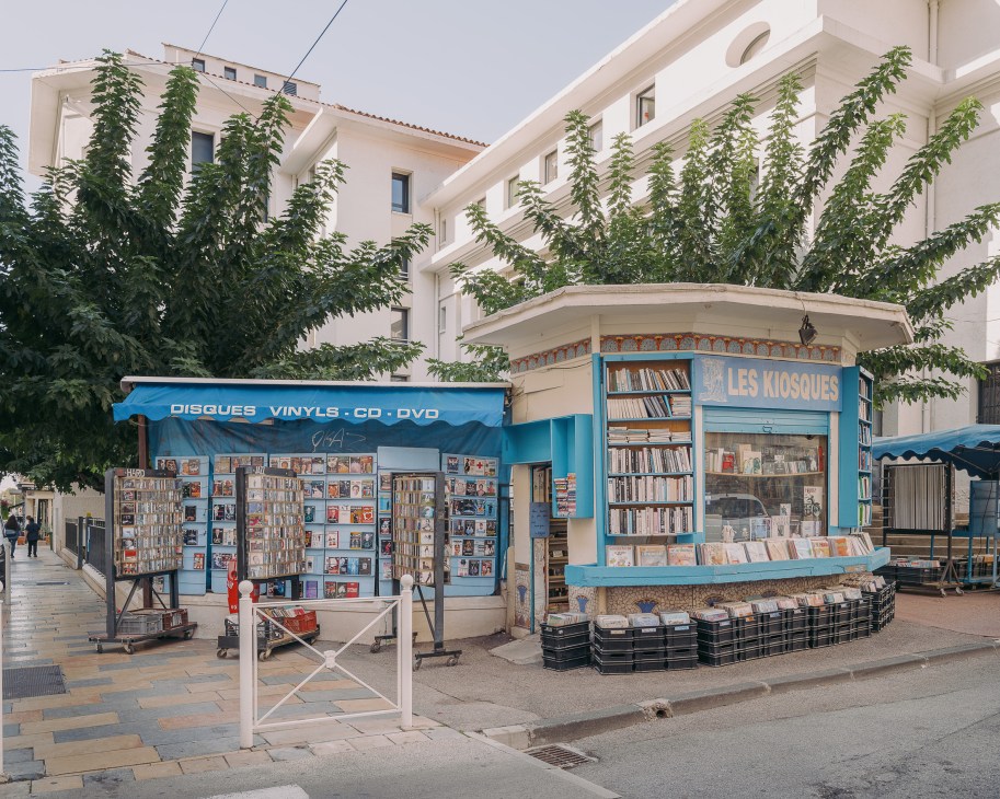 Blue kiosk storefront selling vinyl records, CDs and DVDs on a European street with white buildings.