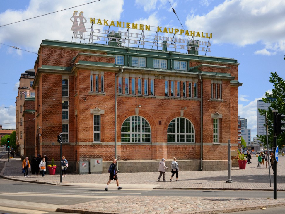 Historic brick building with large arched windows and green roof featuring Hakaniemi Kauppahalli signage