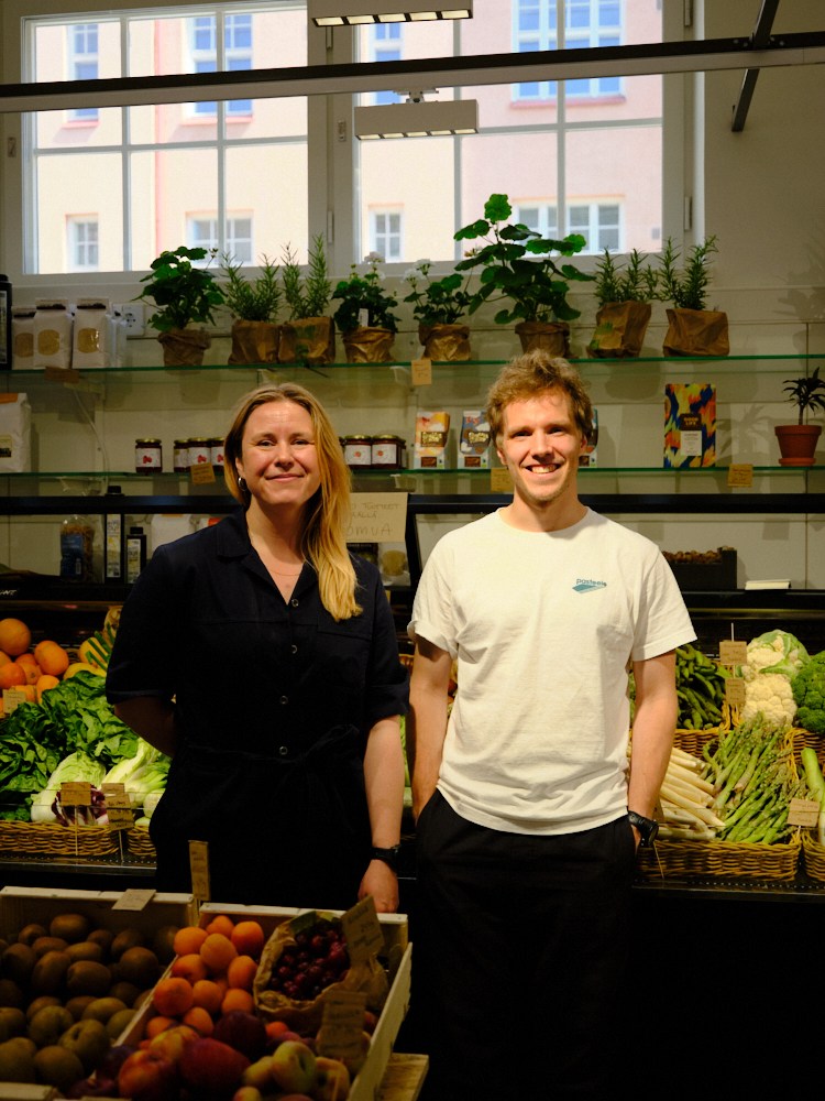 Two people standing in a market hall with fresh produce displays and potted herbs on glass shelves