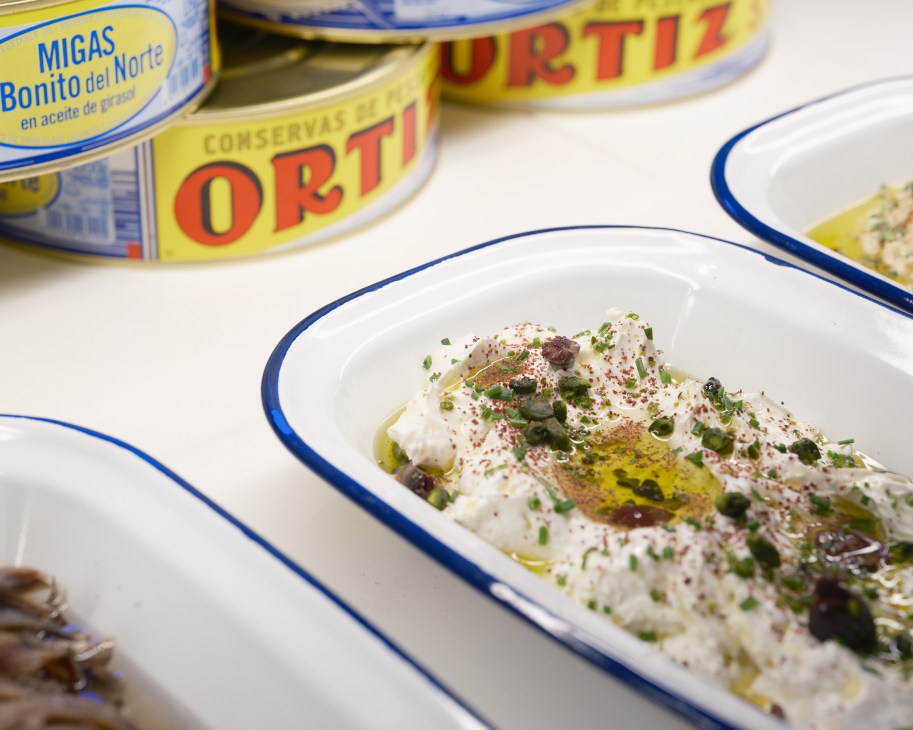 White ceramic dish with creamy spread topped with herbs, olives, and olive oil next to tuna cans