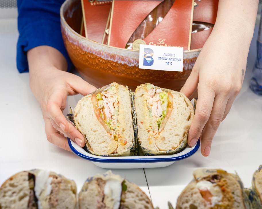 Hands holding a plate with a traditional Niçois pan bagnat sandwich cut in half, showing filling of vegetables and tuna
