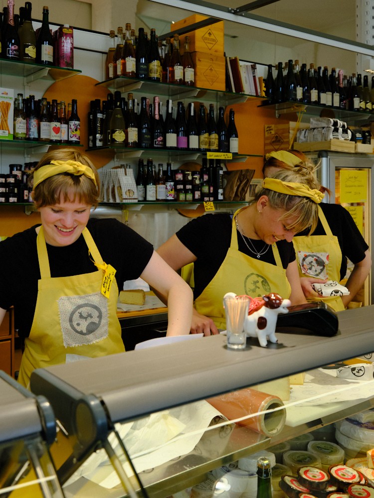 Two staff members in yellow aprons working behind a deli counter with shelves of wine bottles in the background