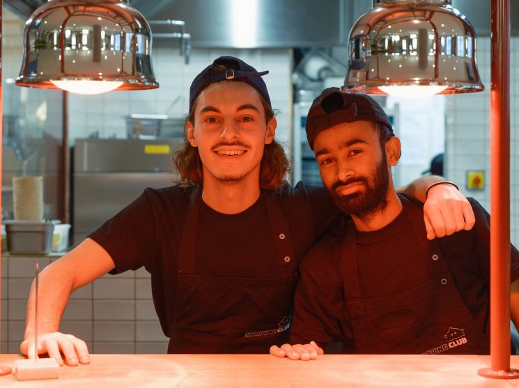 Two smiling staff members in black uniforms posing together behind the counter at Starving Club restaurant