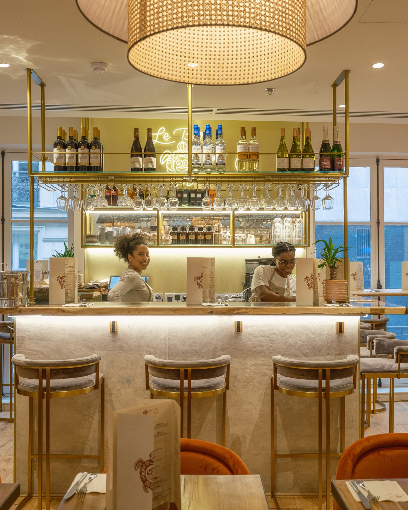 Modern restaurant bar with marble counter, brass fixtures, wine display and two staff members behind counter