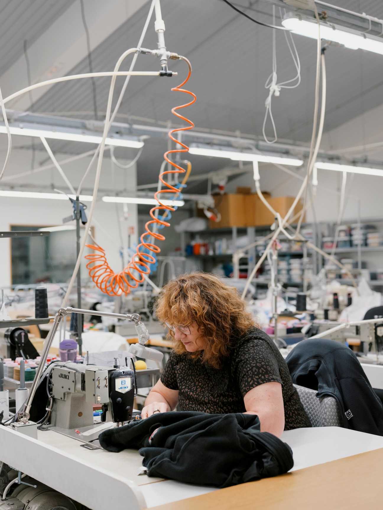 Seamstress working at a sewing machine in a bright workshop with coiled orange cables hanging overhead.