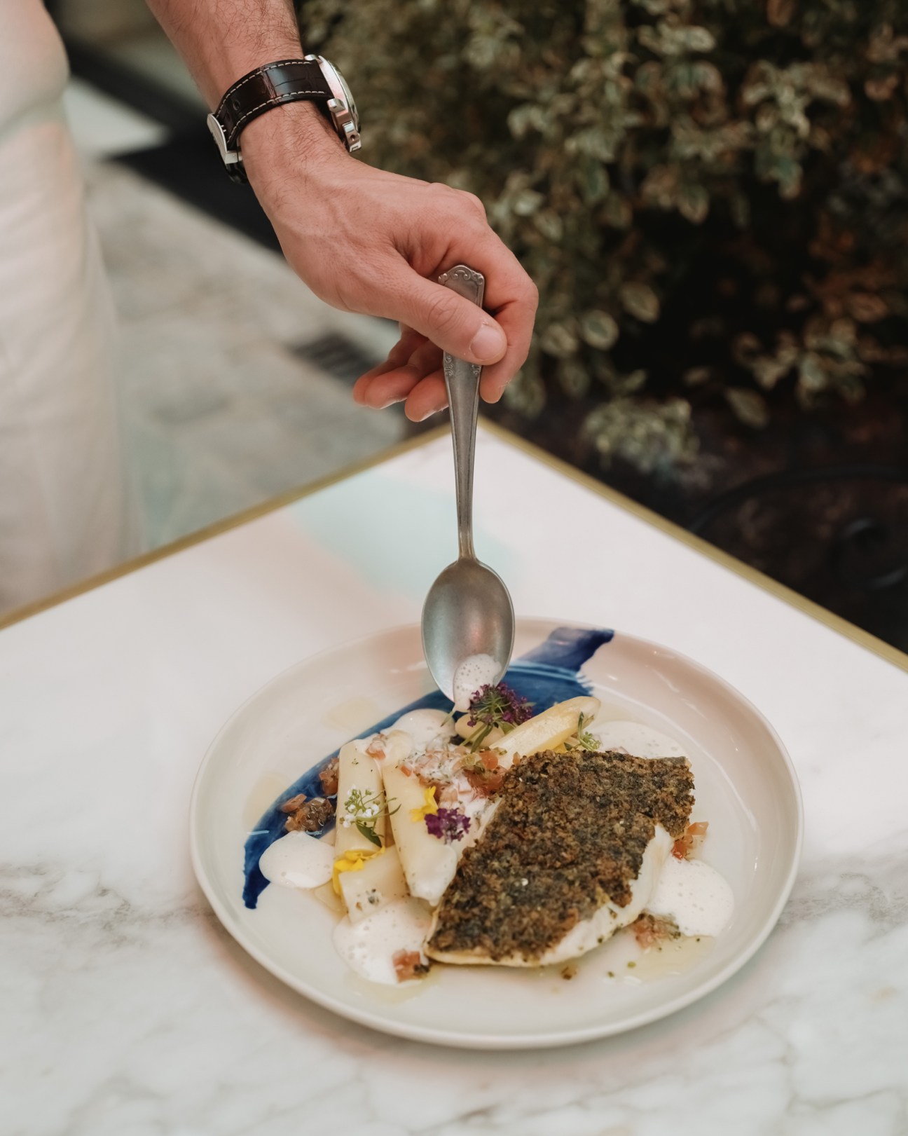 Hand holding spoon over plated sea bass dish with herbs and garnishes at La Chambre Bleue restaurant