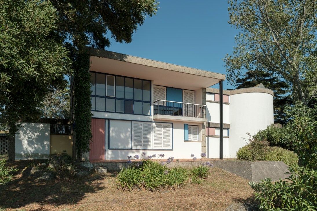 Two-story modernist house with white cylindrical tower, flat roof, large windows and balcony surrounded by trees