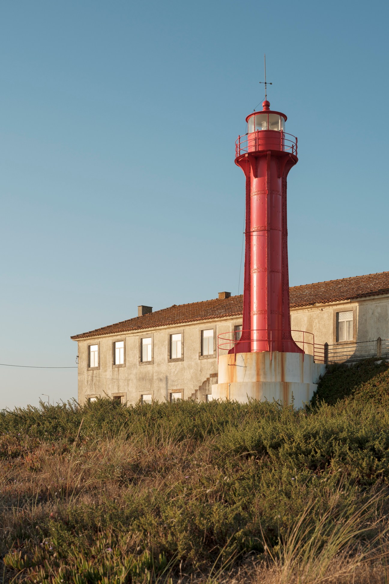 Red lighthouse tower with lantern room and railing, attached to white stone building with clay tile roof